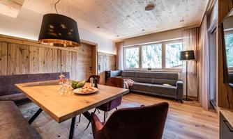 A cozy living space with wooden paneling and large windows. A dining area with a wooden table and comfortable chairs is also visible.