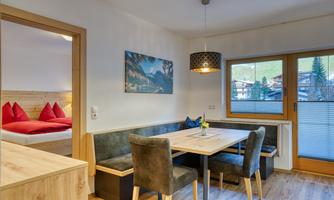 A cozy dining area with a table and chairs. In the background, red cushions are visible in a bedroom.