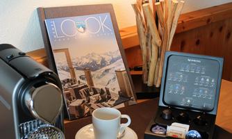 A modern coffee machine is on a table with two cups and a magazine. In the background, a lamp and decorative wood are visible.