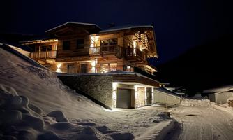 A beautiful chalet in the snow at night, illuminated by warm lights. The surroundings are quiet and the landscape is covered with a layer of fresh snow.