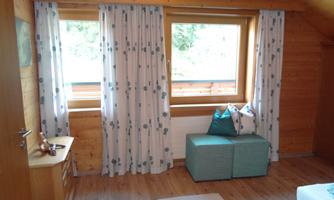 A cozy room with wooden paneling and large windows. The windows are decorated with curtains, and a stool with cushions is placed in the center of the room.