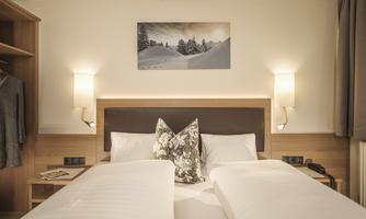 A cozy hotel room with two single beds and modern lamps. A picture of snow-capped mountains hangs on the wall.