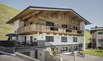 A modern house in alpine style with wooden cladding and balconies. The surroundings are green and inviting, ideal for a peaceful holiday.