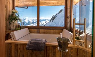 A modern bathroom with a bathtub and large windows that offer a stunning view of snow-capped mountains. The wood-paneled surroundings create a cozy atmosphere.