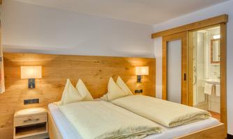 A cozy bedroom with a double bed and wooden accents. The room features soft lighting and an adjacent bathroom option.