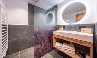 A modern bathroom with dark tiles and an illuminated round mirror. The wooden sink is decorated with fresh towels.