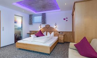 A modern bedroom with a double bed and purple pillows. The walls are light, and the lighting creates a cozy atmosphere.