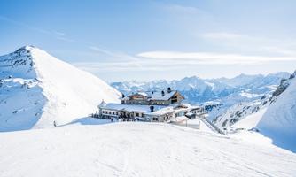 A mountain restaurant in the Alps, surrounded by snow-capped peaks. The sky is clear and bright blue.