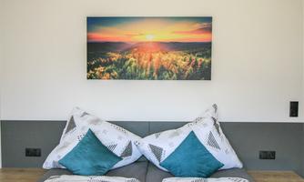 A cozy bedroom with a modern bed and colorful pillows. A picture of a sunset hangs on the wall.