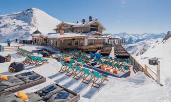 A picturesque mountain hut in the snow with cozy loungers and colorful seating. The surrounding mountains are clear and sunny.
