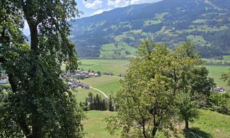 A picturesque landscape with green meadows and mountains in the background. Small houses can be seen in the valley, surrounded by trees.