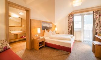 A bright hotel room with two beds and modern furniture. In the background, a bathroom and a window with a view outside can be seen.