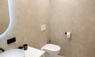 A modern bathroom with bright, neutral tiles. It features a sink, a toilet bowl, and a minimalist decor.