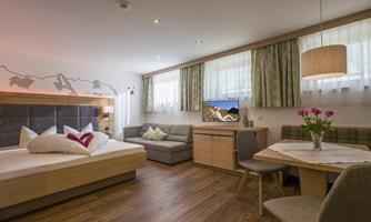 A modern room with a double bed, a cozy seating area, and a dining table. Large windows provide plenty of daylight and create a pleasant atmosphere.