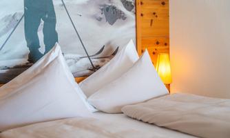 A cozy bedroom with white cushions and a warm lamp. In the background, an inspiring ski motif can be seen.