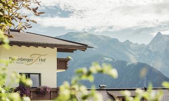 A hotel building with the inscription "Filzvenberger Hof" set against an impressive mountain landscape. The sky is clear and the mountains are surrounded by fog.