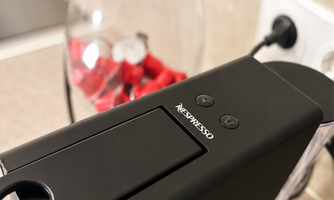 A Nespresso coffee machine in black design is placed on a countertop. In the background, a glass with red fruits can be seen.