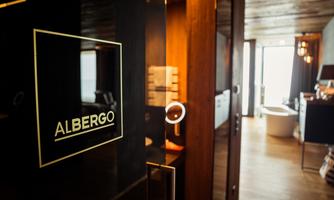 A modern hotel door with the sign "ALBERGO". In the background, a well-designed room with plenty of light can be seen.