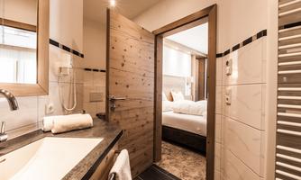 A modern bathroom with a large sink and wooden details. In the background, a bedroom with a cozy bed is visible.