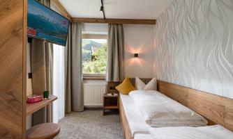 A modern hotel room with wooden paneling and a large window. The room features a television, a cozy sofa, and a decorative pillow.