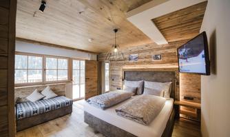 A modern bedroom with wood paneling and large windows. It features a comfortable double bed and a cozy seating area.