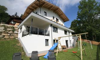 A modern house with white walls and a large balcony. In the foreground, there is a swing and a slide on a green lawn.
