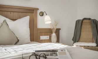 A cozy bedroom with a wooden bed and a reading light. On the bed are a pair of glasses and a magazine, while a wicker chair stands beside it.