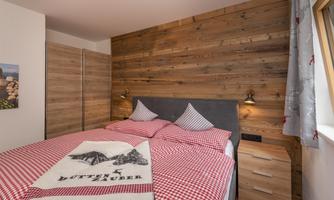 A cozy bedroom with a wooden wall design and a large double bed. The red-and-white checkered bed linen and the bedside table give the room an inviting atmosphere.