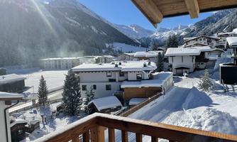 A picturesque winter landscape with snow-covered houses and mountains in the background. The sky is clear and the sun is shining.