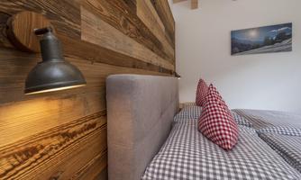 A cozy bedroom with a wood wall design and two beds. The pillows are checked, and there is a warmly glowing wall lamp.