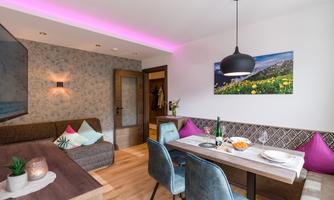 A modern living room with a cozy sofa, a dining table, and colorful cushions. The walls are tastefully decorated, and soft lighting creates an inviting atmosphere.