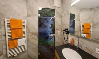 A modern bathroom with marble-patterned walls and a round sink. Towels in bright orange and a shower cabin with a natural motif complement the space.
