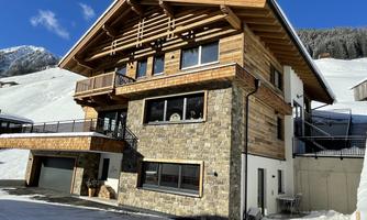 A modern chalet made of wood and stone elements in a snowy mountainous landscape. The clear sky and surrounding mountains give the scene a picturesque atmosphere.