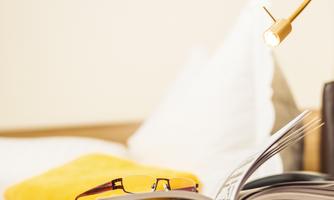 A cozy bed with a pillow in the background and an open book in the foreground. A pair of glasses and a yellow towel complement the relaxed atmosphere.