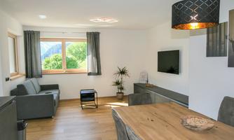A modern living room with a cozy sofa, a dining table, and a television. Large windows provide a view of the greenery.