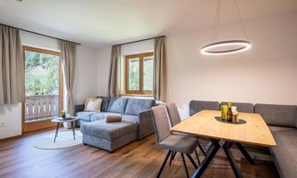 A modern living room with a grey sofa and a wooden dining table. Large windows let in plenty of light and provide a view outside.