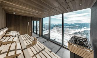 A modern sauna with wooden interiors and large windows. The view shows the snow-covered mountains and the clear sky.