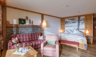 A cozy hotel room with wooden elements and a warm atmosphere. There is a seating area with a red sofa and a view of a large bed.