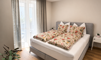 A cozy bedroom with a large bed and colorful bedspreads. The windows let in a lot of light and plants decorate the space.
