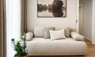 A modern living room with a bright sofa and a decorative cushion. In the background, a piece of art hangs above a green plant in a basket.