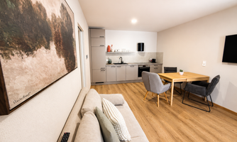 A modern living kitchen with a sofa, dining table, and chairs. The walls are light and there is a large piece of artwork.