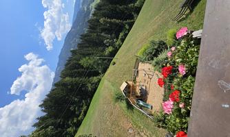 A playground area with a wooden playhouse and colorful flowers in the foreground. In the background, green meadows and wooded mountains stretch under a blue sky.