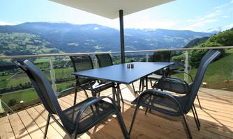 A modern table with chairs on a terrace overlooking the mountains. The sky is clear and the landscape is green and inviting.