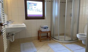 A bright bathroom with a shower cubicle, sink, and toilet. Clean towels are placed on a wooden chair, and the room has tiled floors.